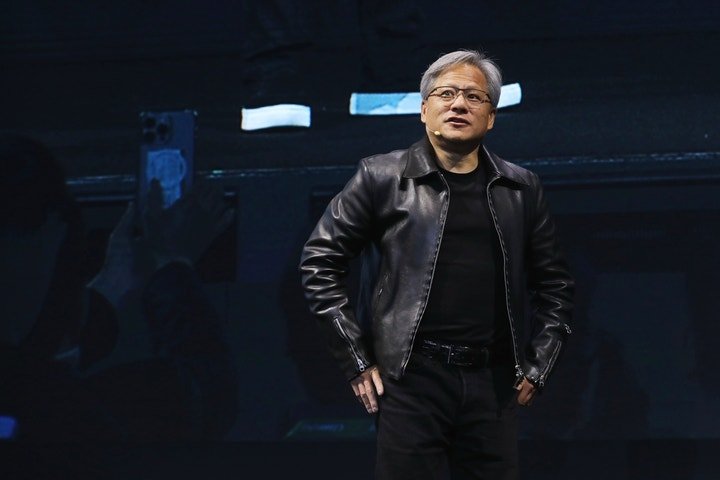 A person wearing a black leather jacket and black shirt stands confidently on stage, gesturing slightly with one hand while looking towards the audience.