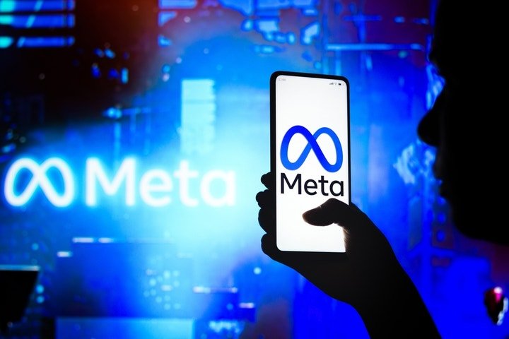 "Silhouette of a person holding a smartphone displaying the Meta logo, with a blurred background featuring the Meta logo in blue lighting."