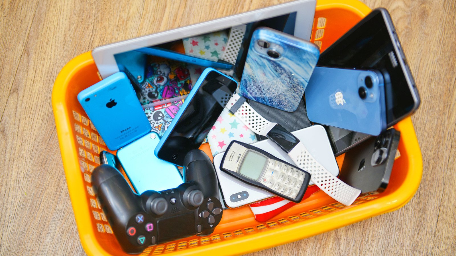 A cluttered orange basket filled with various electronic devices, including smartphones, a tablet, a gaming controller, and a vintage mobile phone, all resting on a wooden surface.