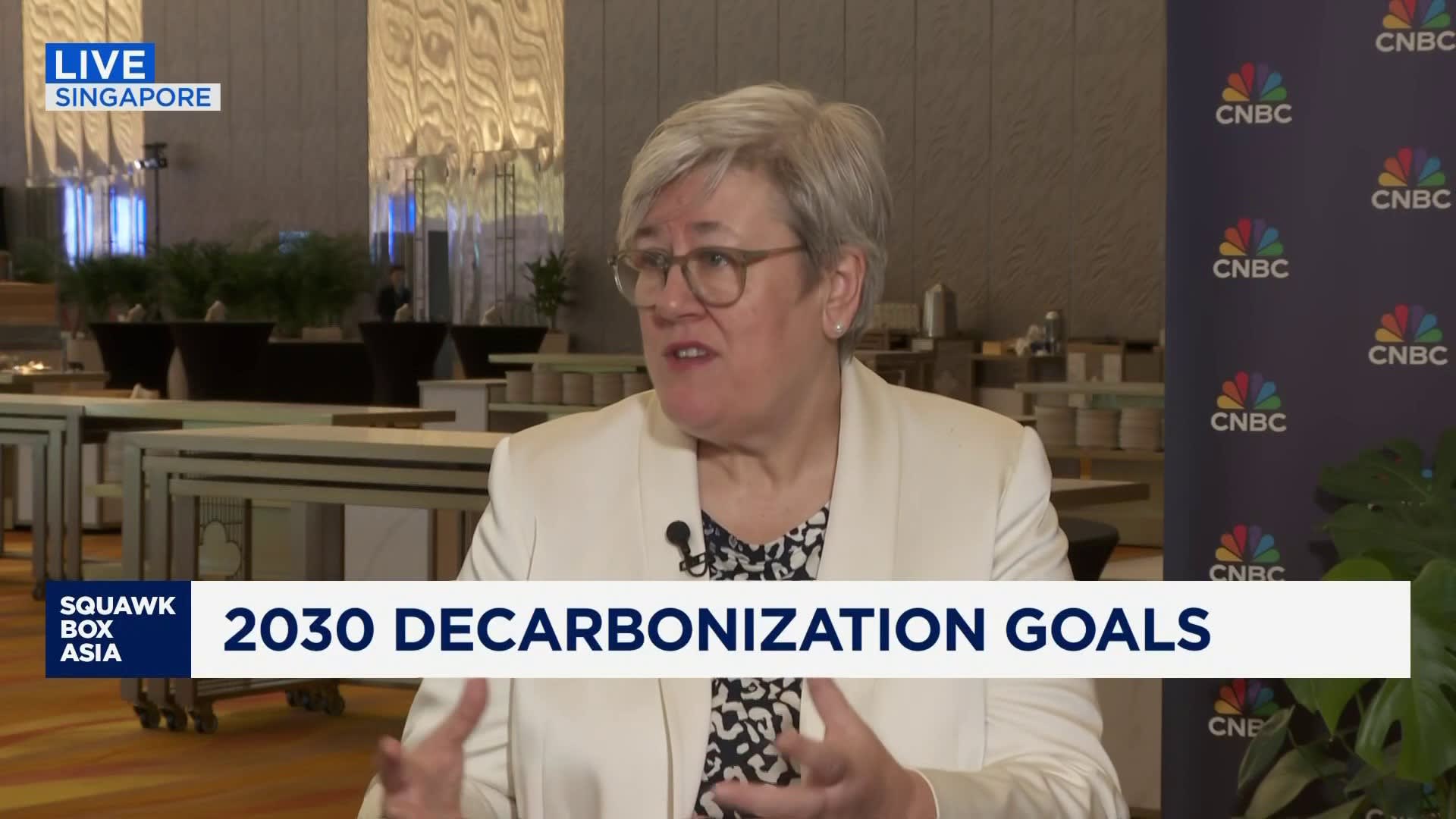 "Interview segment featuring a woman with short gray hair and glasses, speaking animatedly about decarbonization goals in a modern indoor setting with plants and tables in the background. The text overlay includes 'LIVE SINGAPORE' and '2030 DECARBONIZATION GOALS.'"