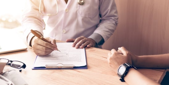 "A doctor in a white coat writing on a clipboard while a patient with a wristwatch sits across the table, hands resting on the surface."