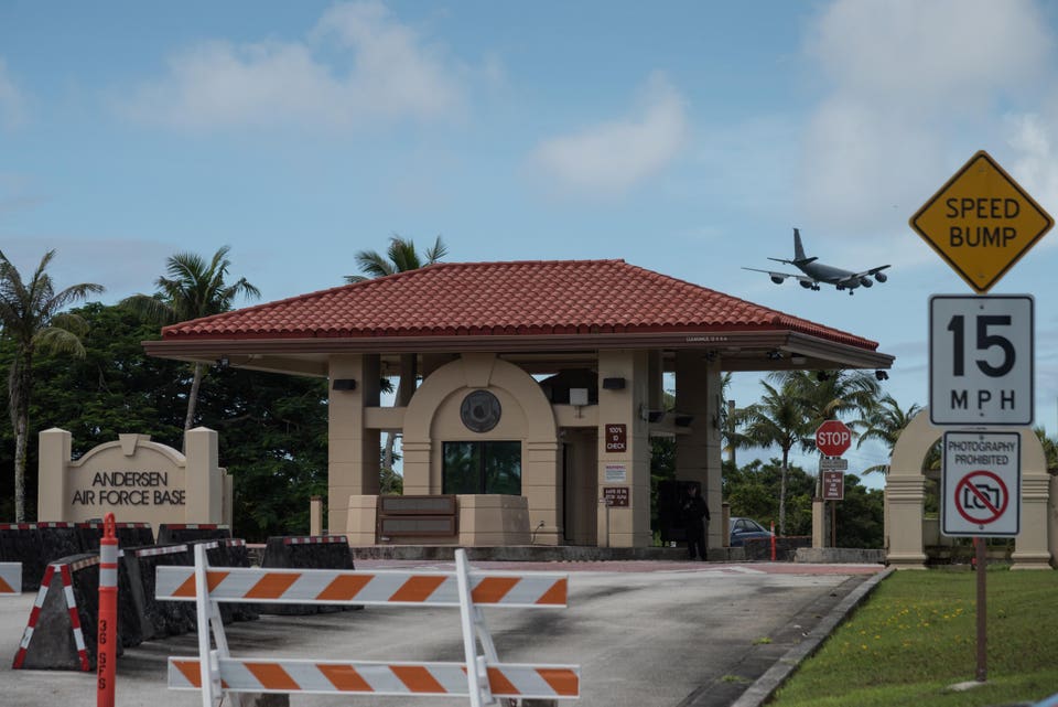 Entrance to Andersen Air Force Base with a security checkpoint, palm trees in the background, and a military aircraft flying overhead. A sign indicates a speed bump and another warns against photography. Traffic barriers are visible in front of the entrance.