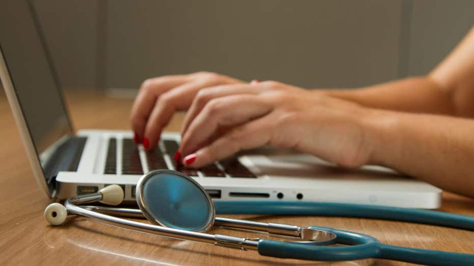 "Hands with red nail polish typing on a laptop keyboard, with a stethoscope resting on a wooden surface nearby."