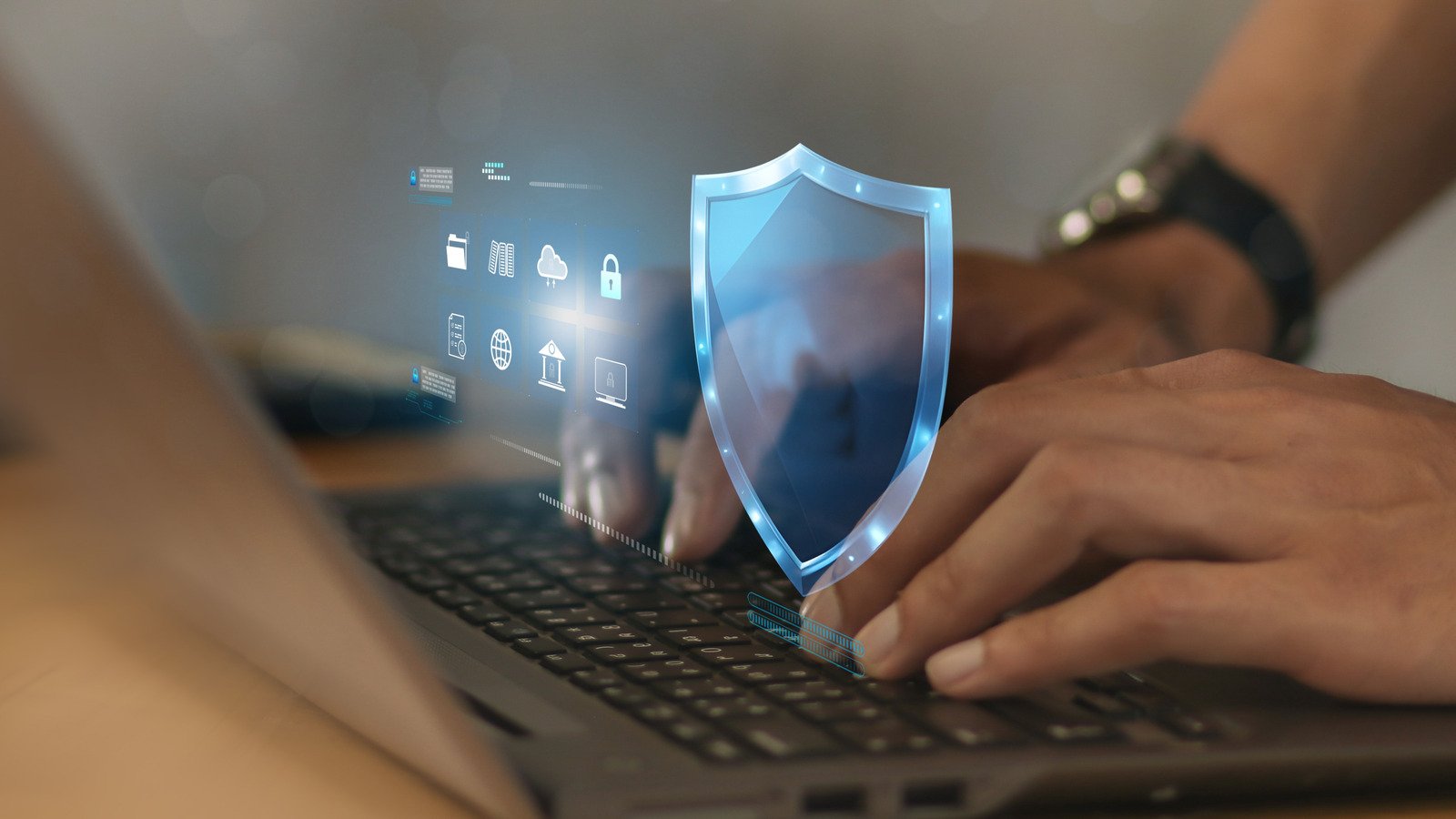 "Close-up of hands typing on a laptop, with a digital shield icon and various security-related symbols displayed on the screen."
