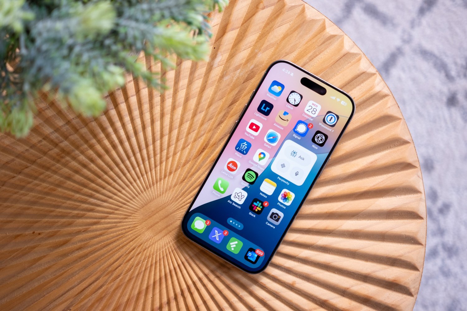 "Smartphone displaying various app icons on the screen, placed on a textured wooden surface with a circular pattern. A small green plant is partially visible in the background."
