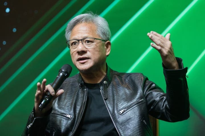 "Person in a black leather jacket speaking into a microphone while gesturing with their hands, set against a green background with diagonal lines."