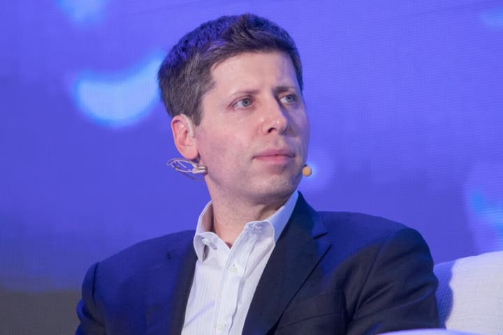 A man with short, light brown hair wearing a dark blazer and a light-colored shirt, seated and looking thoughtfully to the side, with a microphone attached to his ear, against a blue background.