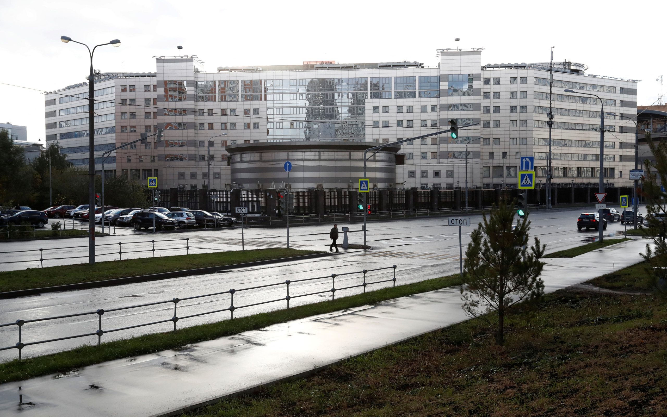 Modern building with large glass windows and a cylindrical section, situated across a wet road lined with streetlights and traffic signs. A person walks on the sidewalk near parked cars and greenery.