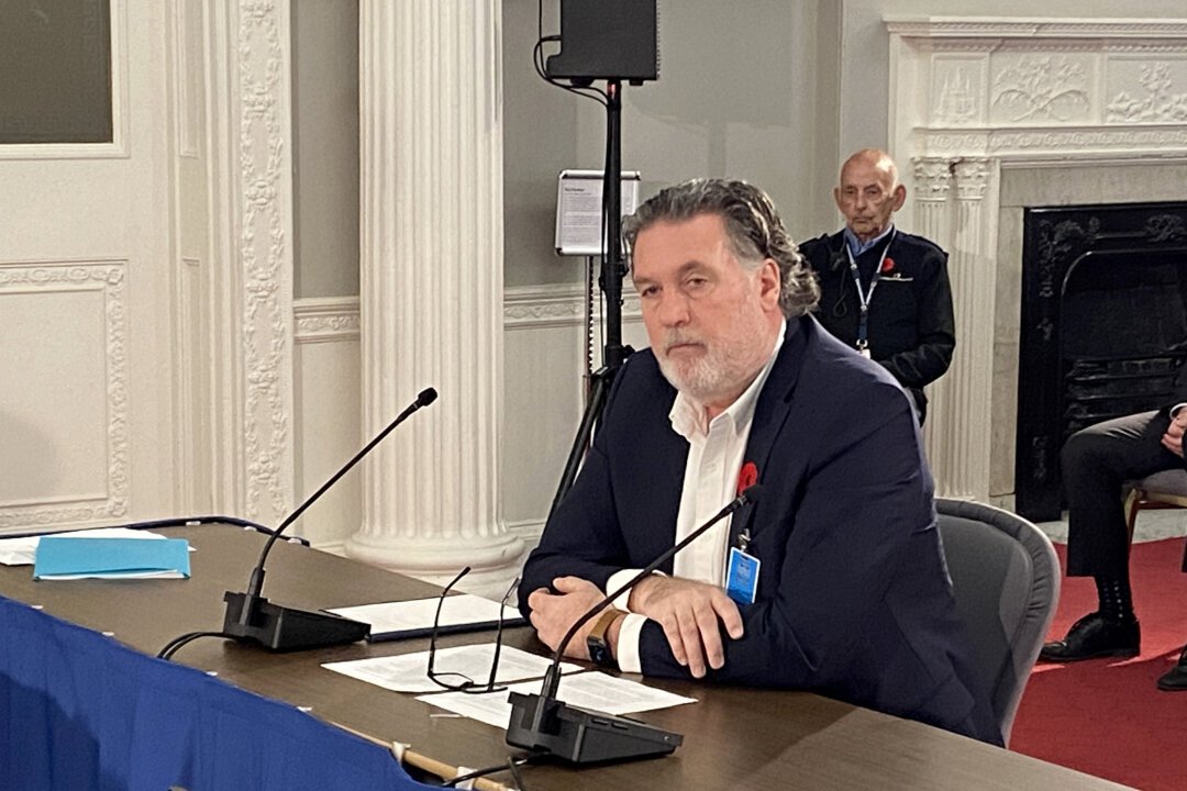 A man with a beard and dark hair, wearing a dark suit and white shirt with a red poppy, sits at a conference table with microphones, looking at the camera. Ornate white walls and a fireplace are visible in the background.