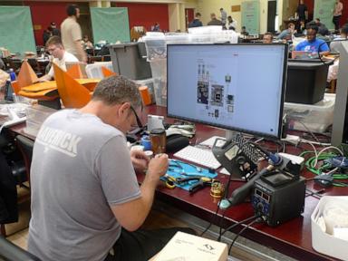 Man soldering electronics at a desk with a monitor displaying a circuit design in a workshop.