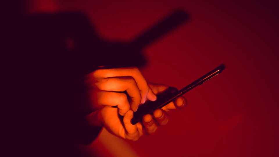 Hands typing on a smartphone, illuminated by the screen's glow against a dark red background.
