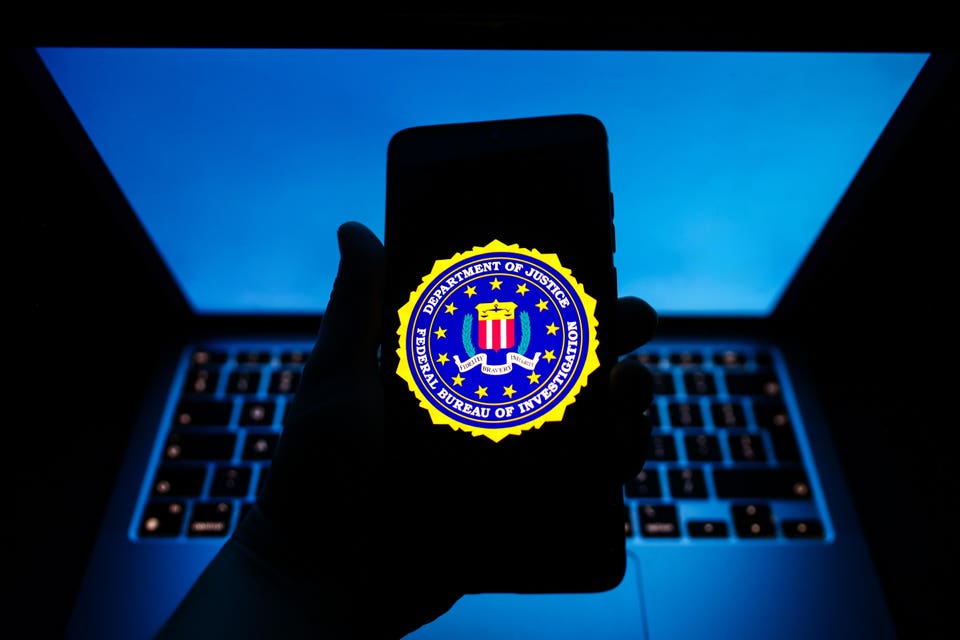 "Hand holding a smartphone displaying the FBI seal, with a backlit laptop keyboard in the blue-lit background."