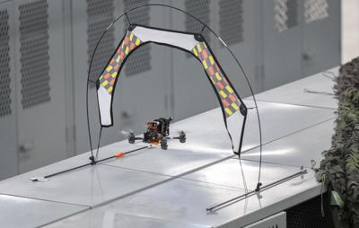 Small drone flying through a white archway with orange, black, and yellow checkered panels, set up on an indoor surface.