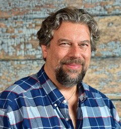 Smiling man with curly graying hair and a beard, wearing a blue and white plaid shirt.