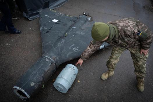 Ukrainian soldier in camouflage examines the wreckage of a large dark drone or missile, with a detached cylindrical part lying on the wet ground.