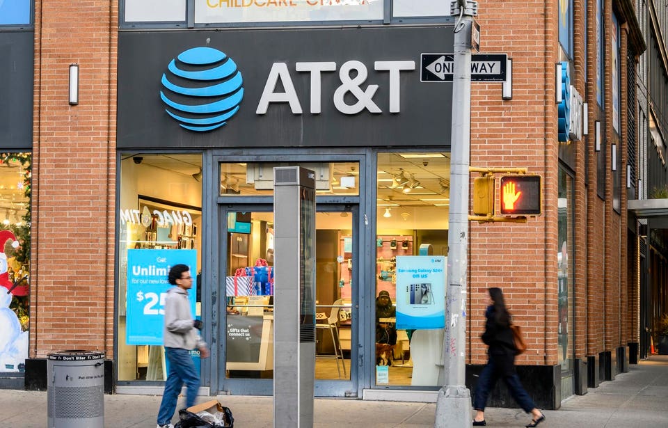 AT&T store exterior with its blue globe logo on a brick building, featuring a One Way sign and a red pedestrian signal, with people walking on the sidewalk.