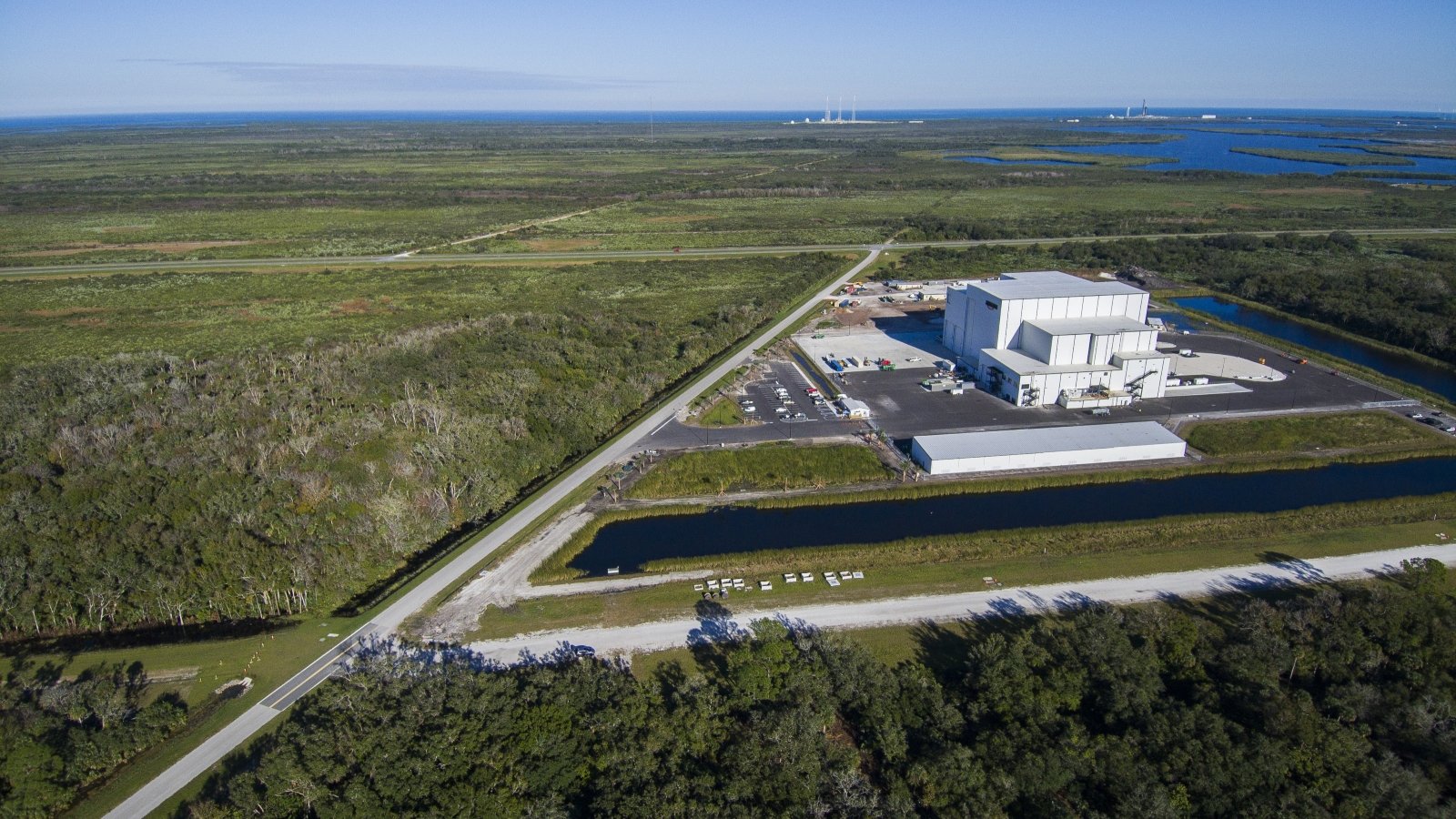 Aerial view of a large white facility building with an access road, surrounded by a vast green landscape and waterways. The ocean and distant launch towers are visible on the horizon.