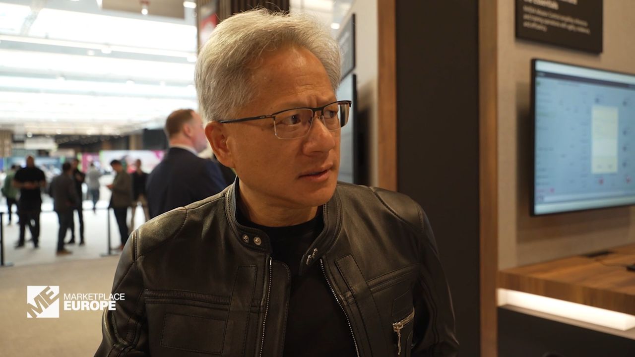 Jensen Huang, CEO of Nvidia, wearing a black leather jacket and glasses, looks to the right at the Marketplace Europe event, with blurred attendees and a screen in the background.