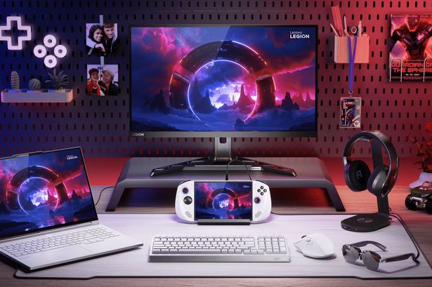 Lenovo Legion gaming desk setup with a monitor, laptop, and handheld device displaying a futuristic game scene. A white keyboard, mouse, and black headphones are on the desk, backed by a pegboard wall with gaming decor.
