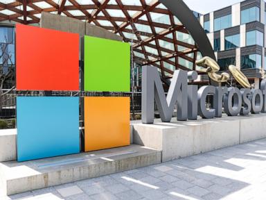 The Microsoft logo with four colored squares and the wordmark in large grey letters on a concrete base, set outdoors in front of modern buildings.