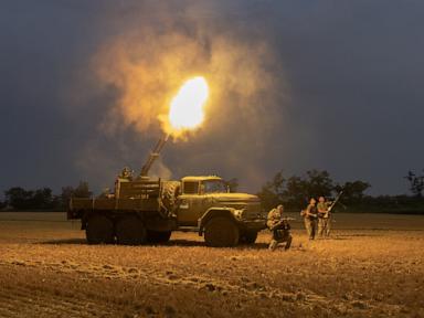 A military multiple rocket launcher truck fires, emitting a large fiery blast into the night sky, while soldiers stand in a field.