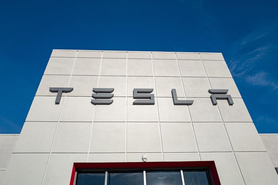 Tesla logo in grey letters on the light-colored, paneled facade of a modern building, beneath a clear blue sky.