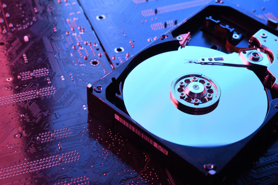 Opened hard drive showing its reflective data platter and read/write head, positioned on a dark circuit board with red and blue lighting.