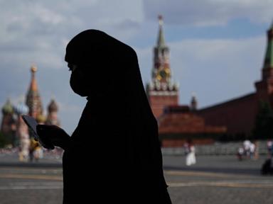 "Silhouetted person wearing a head covering and face mask, looking at a mobile phone with St. Basil's Cathedral and Kremlin towers in the background."