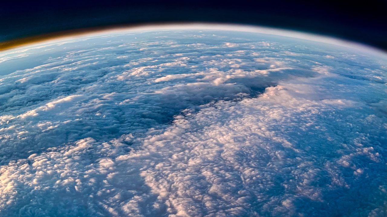 Aerial view of Earth showing a vast layer of white and gray clouds, with the planet's curvature visible against the dark atmosphere and a subtle golden horizon.