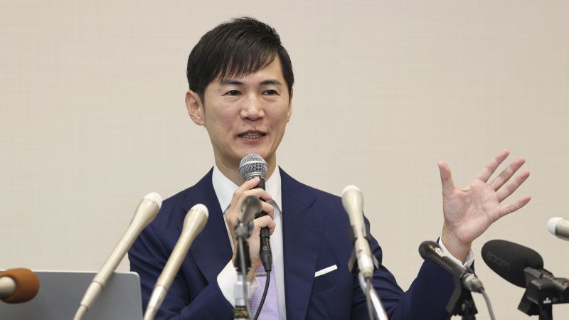 Man in a suit speaking into a microphone and gesturing with his left hand at a press conference.