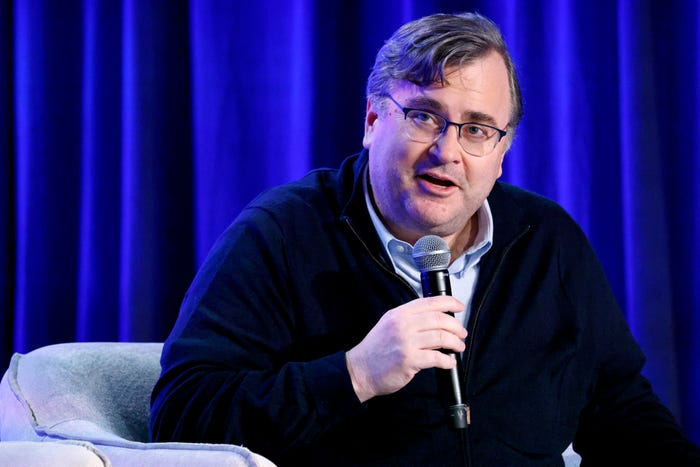 Man in glasses and a dark blue sweater speaking into a microphone against a blue curtain.