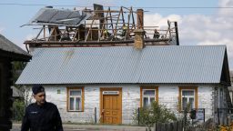 White building with exposed roof rafters and workers on the roof, with a person in a dark uniform in the foreground.