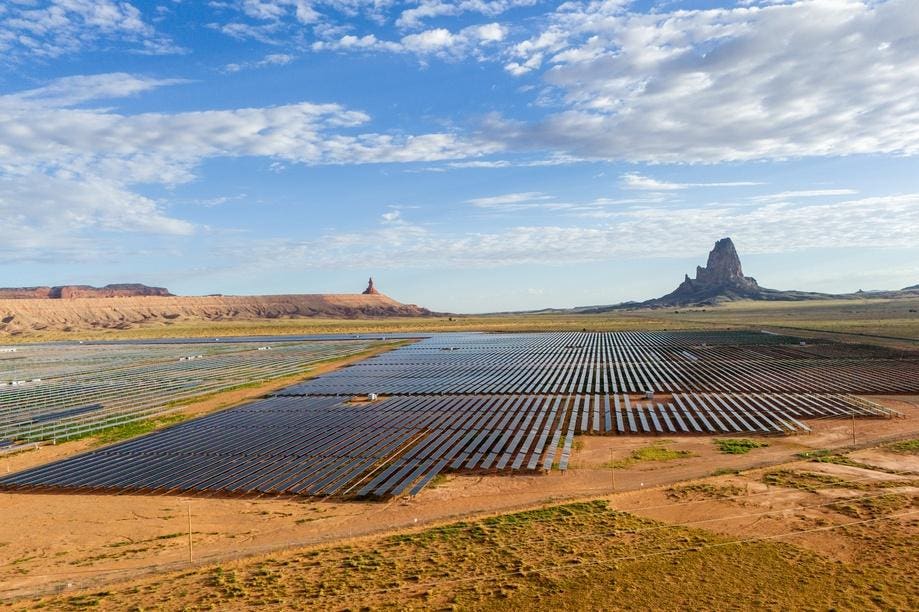"Rows of solar panels in a desert landscape, with a large, distinctive rock formation to the right and a mesa in the distance to the left, under a blue sky with scattered clouds."