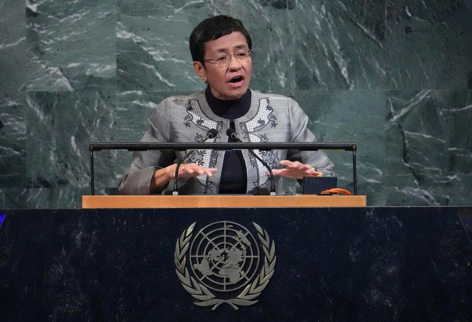 Woman with short dark hair and glasses speaking at a podium with the United Nations logo, gesturing with her hands, against a green marble wall.