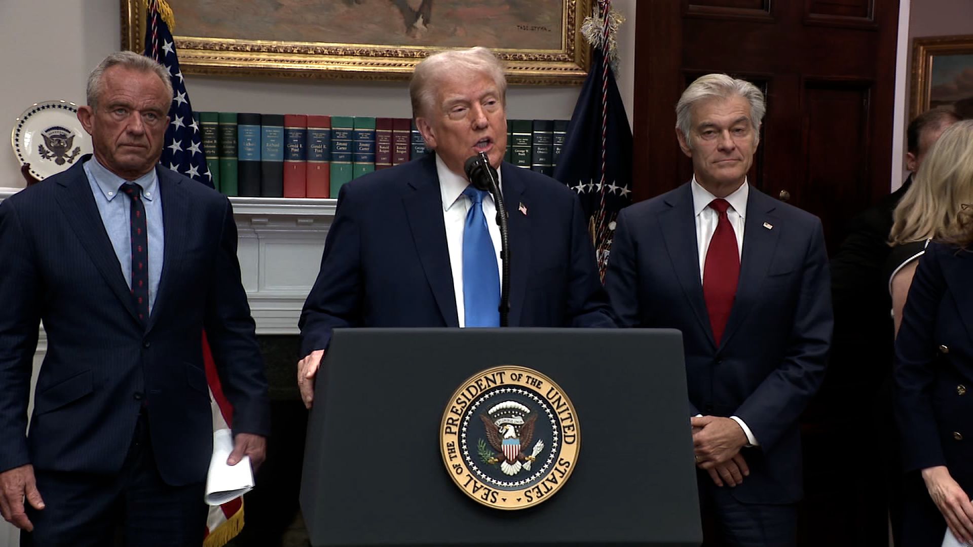 Donald Trump speaking at a podium with the Presidential Seal, flanked by Robert F. Kennedy Jr. and Dr. Mehmet Oz.