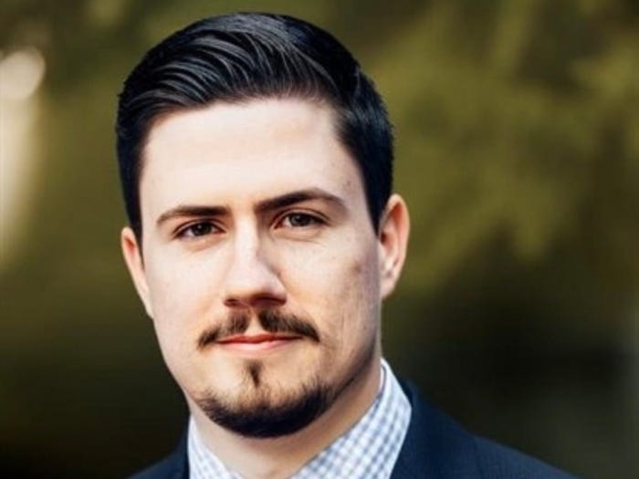 "Professional headshot of a man with dark hair, a mustache, and a goatee, wearing a suit jacket and a plaid shirt."