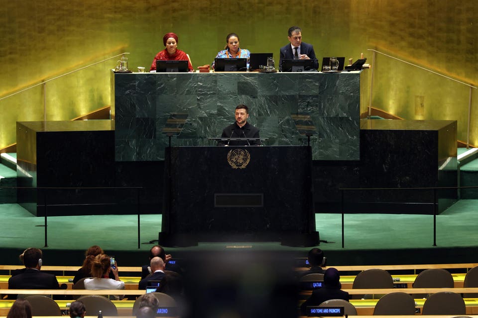 Volodymyr Zelenskyy speaking at a podium with the UN emblem, with three officials seated at a table behind him and an audience in the foreground.