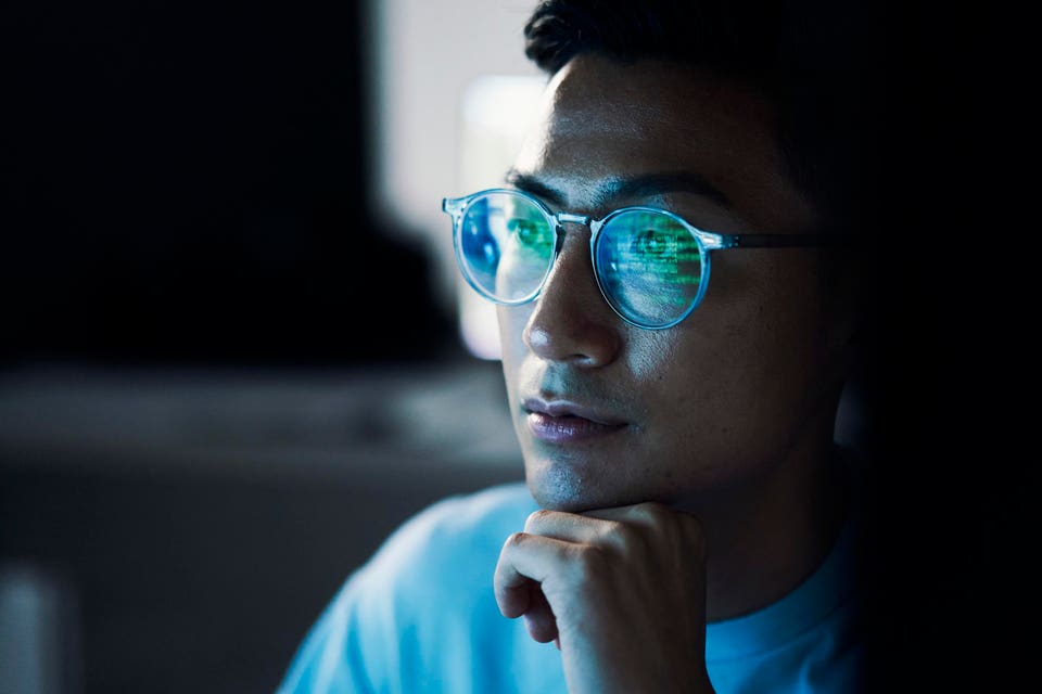 Focused young man in glasses, illuminated by a screen with green digital code reflected in the lenses.