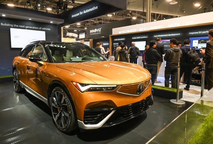An orange SUV with a black roof on display at an automotive technology exhibition, with an "Amazon for Automotive" sign and attendees in the background.