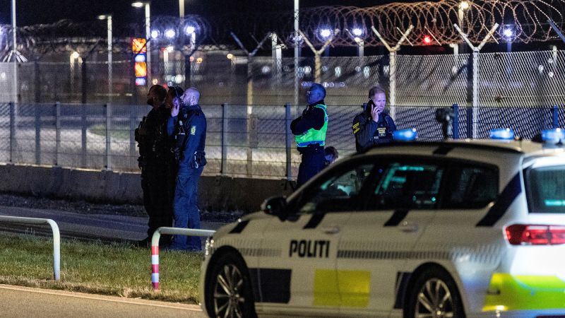 Danish police officers and a police car with blue lights on, at night near a security fence topped with barbed wire.