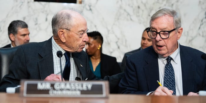 Senator Chuck Grassley, identified by his nameplate, speaking into a microphone at a hearing, with another man in a suit holding a pencil nearby.