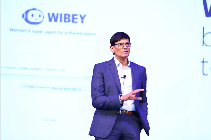 Man in a blue suit and glasses presenting on stage with the "WIBEY" logo and "Walmart's super agent for software teams" displayed on a screen behind him.