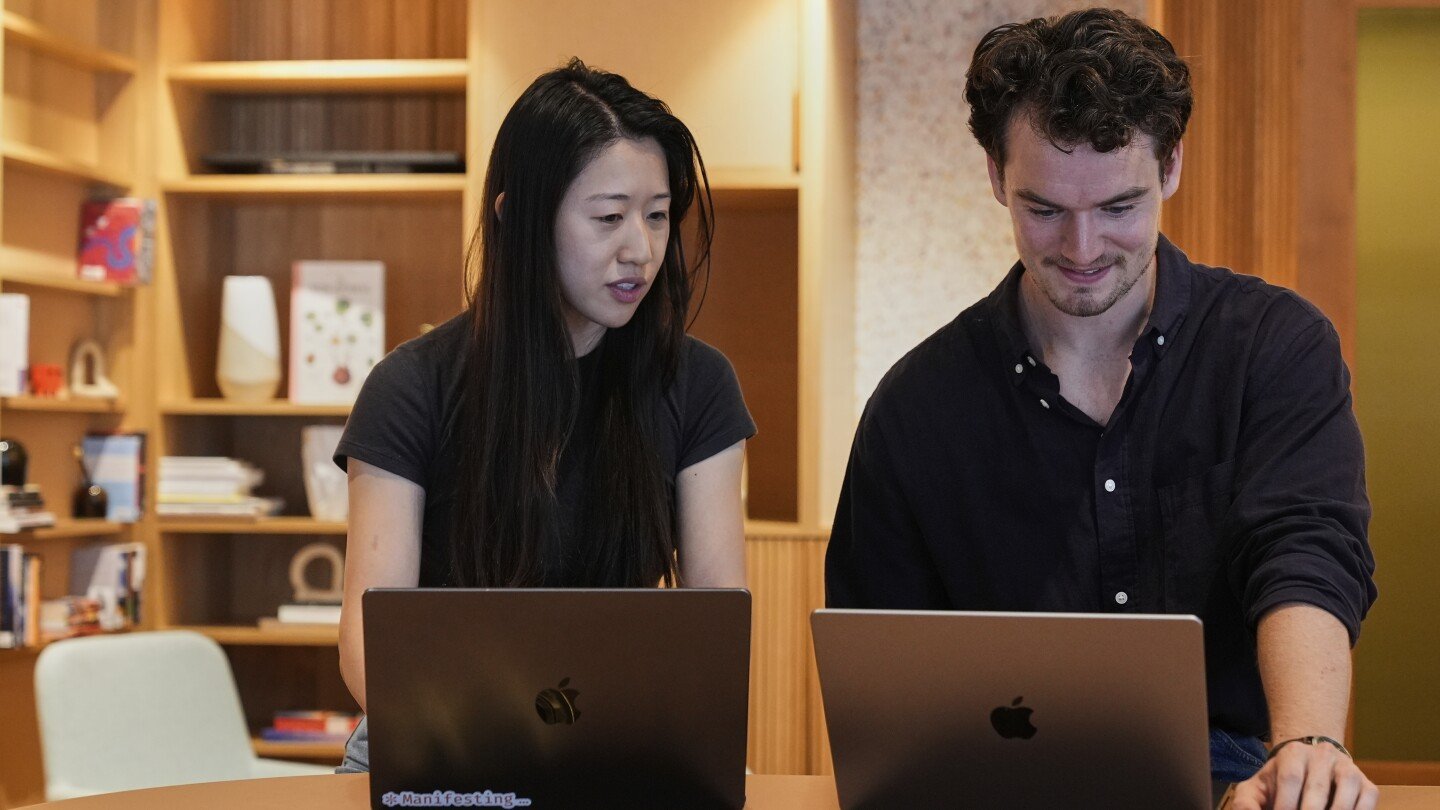 A woman and a man working on laptops at a table, with bookshelves in the background.