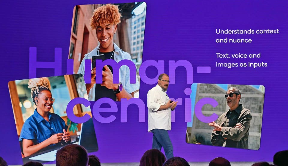 A presenter stands before a large purple screen displaying the words 'Human-centric', overlaid with three tilted images of diverse individuals using technology. The screen also shows text: 'Understands context and nuance' and 'Text, voice and images as inputs'.