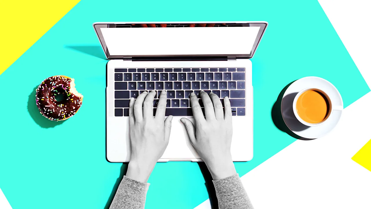 "Hands typing on a laptop with a blank screen, a bitten chocolate donut, and a cup of coffee on a colorful background."