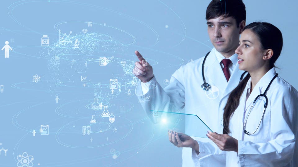 Two medical professionals in lab coats, a man pointing at a holographic display of healthcare data icons, and a woman holding a transparent tablet.