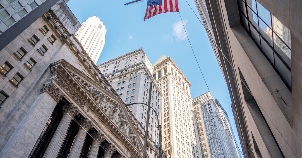 Facade of the New York Stock Exchange building with classical columns and ornate carvings, surrounded by tall urban buildings, and an American flag flying against a blue sky.