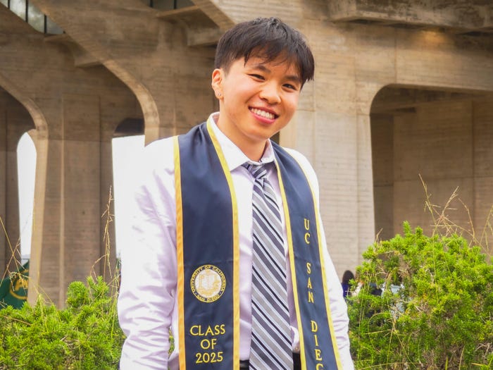 Smiling student in a white shirt and striped tie, wearing a navy UC San Diego stole with "Class of 2025" and the university seal, against a concrete building and green plants.