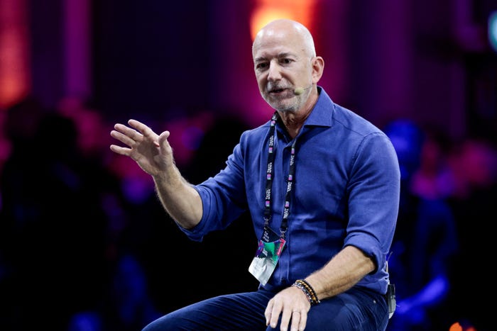 Jeff Bezos, bald and wearing a blue collared shirt, speaks into a microphone and gestures with his right hand at a public event.