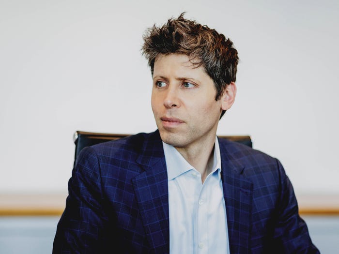 Man with tousled brown hair and light eyes wearing a blue plaid suit jacket, looking to the right.